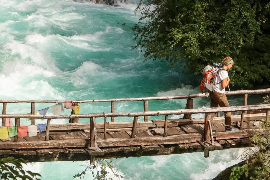 Trekker crossing wooden bridge over turquoise glacial river in Upper Dolpo